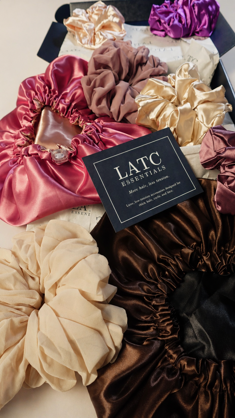 Collection of colorful satin scrunchies  and bonnets with a LATC Essentials box in the center.